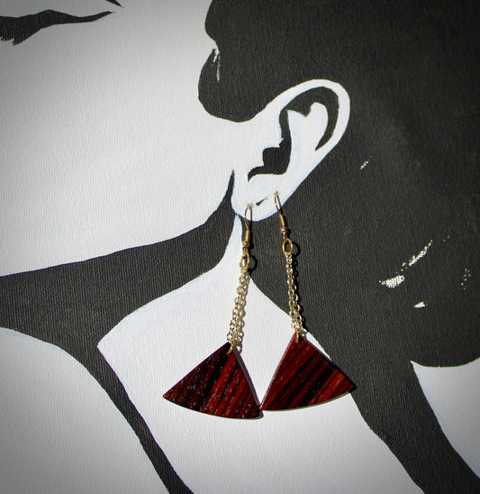 A pair of fan-shaped cocobolo wood earrings with gold chains, displayed against a black background with a silhouette of a woman's face.