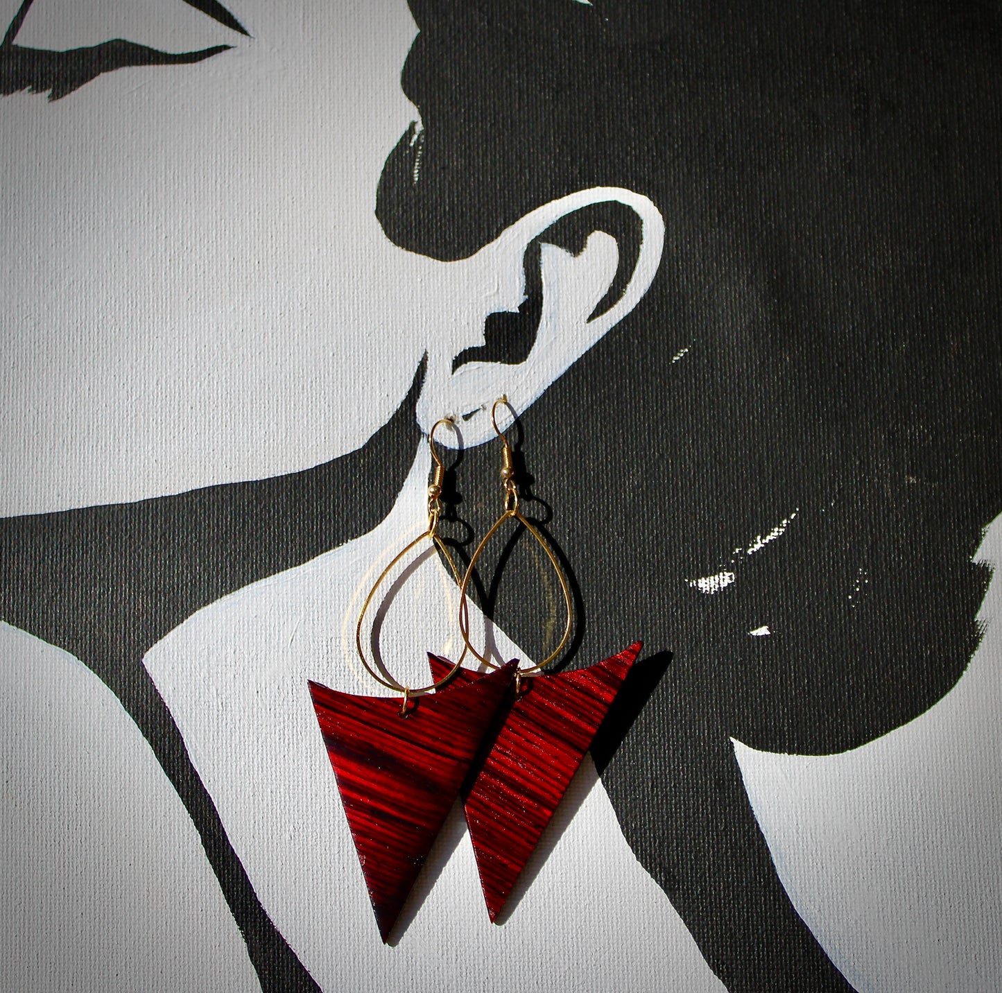 A pair of gold teardrop hoop earrings with red-brown cocobolo wood accents, displayed against a black background with a silhouette of a woman's face.