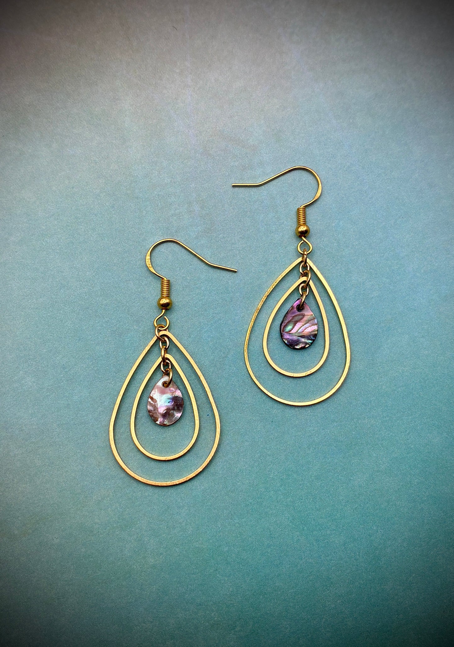 Gold teardrop-shaped earrings with a central abalone pearl on a teal background