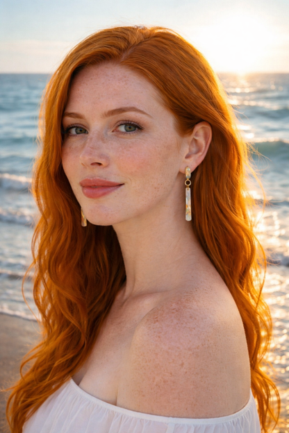 Woman with red hair standing on a beach with ocean in the background wearing gold earrings with abalone pearl drops from Martin Guitar factory materials.