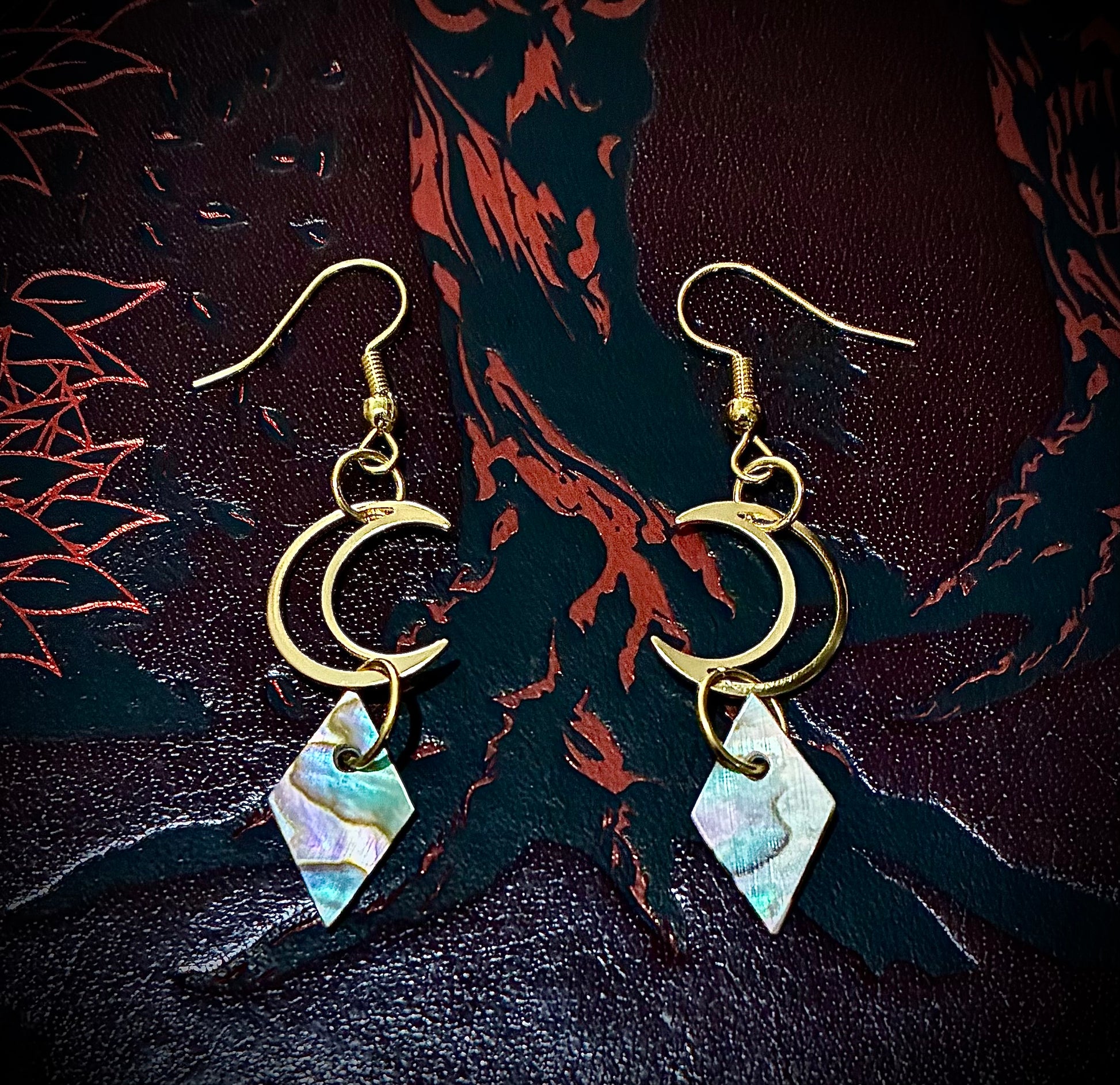 Gold earrings with crescent moon and diamond-shaped Martin guitar abalone pearls on a dark textured background