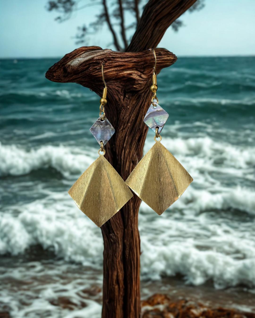 Gold geometric earrings with purple stones on a wooden stand against a beach background.