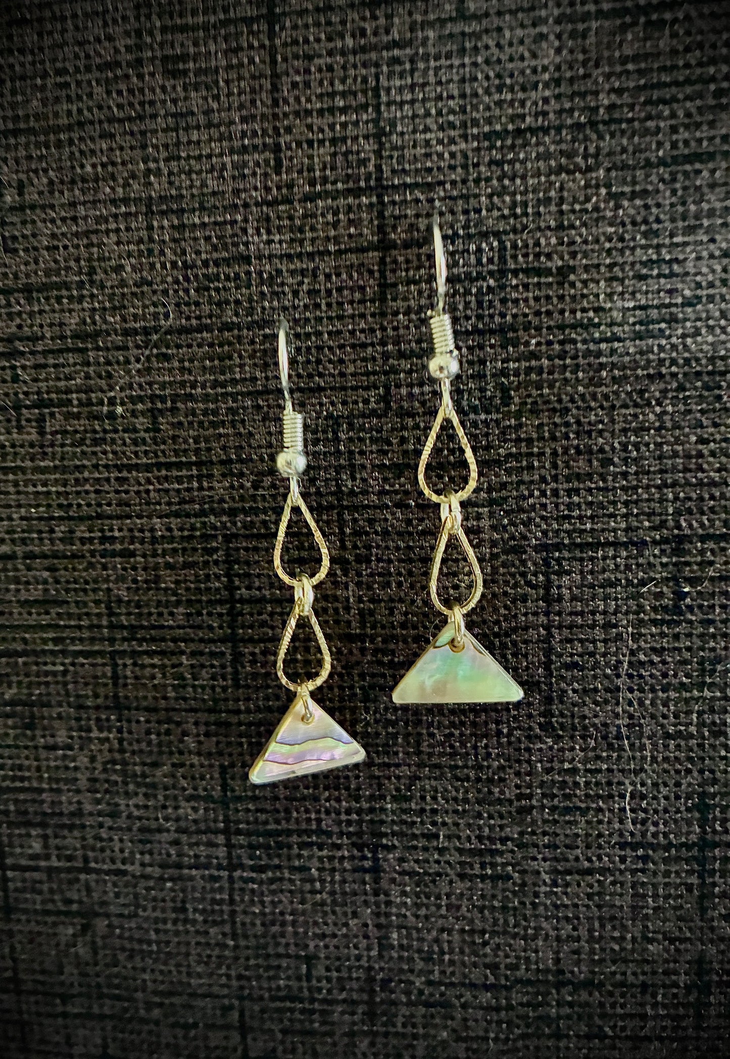 Gold drop earrings in shape of two tears with triangle abalone pearl at bottom against black backdrop.