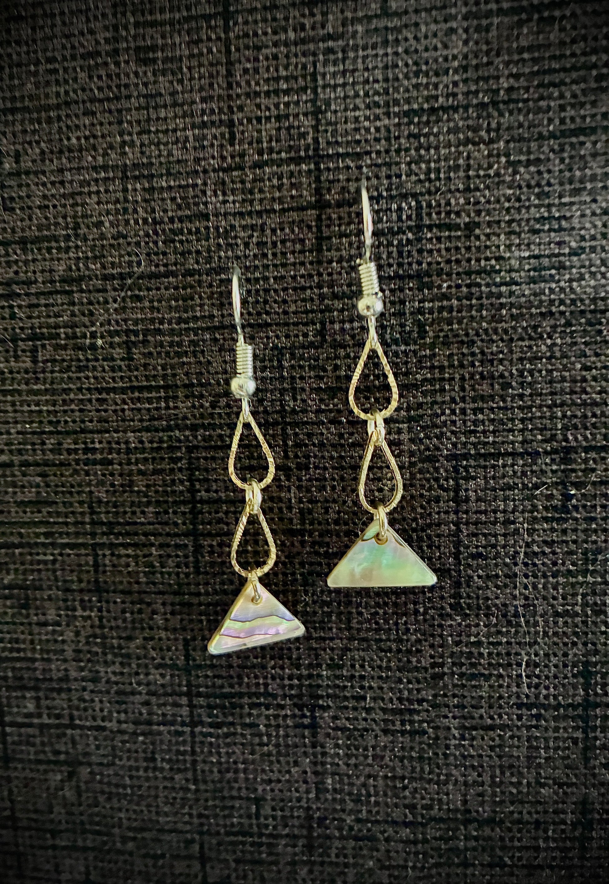 Gold drop earrings in shape of two tears with triangle abalone pearl at bottom against black backdrop.