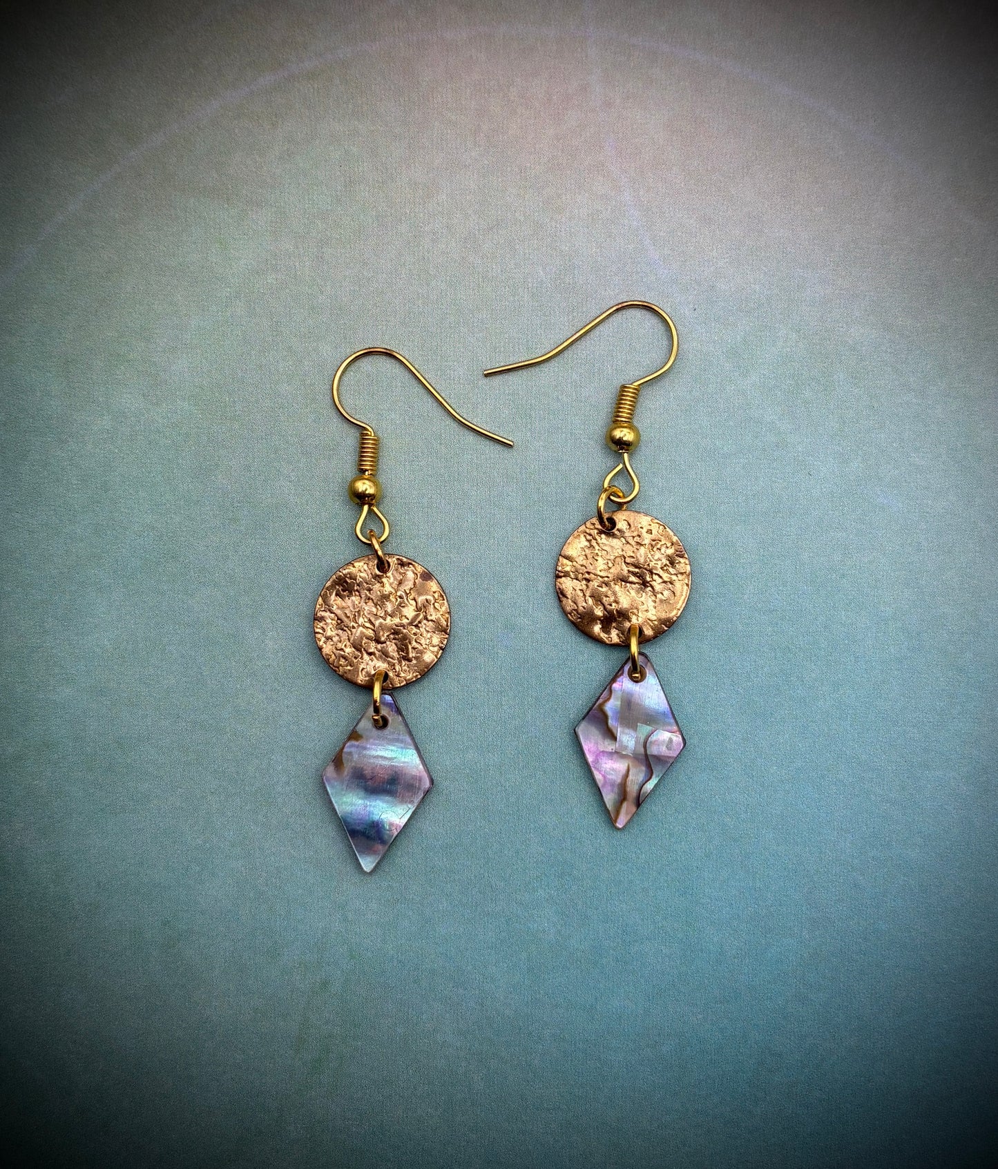 Gold earrings with diamond-shaped abalone pearl drops on a light blue background