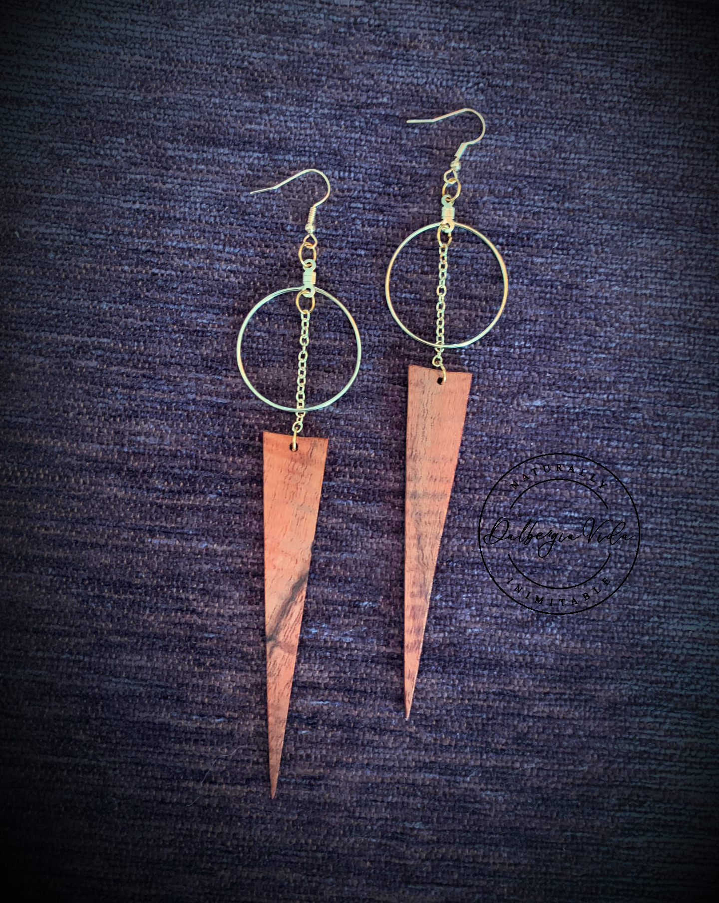 Gold hoop earrings with connecting gold chain to wild grain rosewood spear shaped wood pieces against purple carpet background. Martin guitar factory.